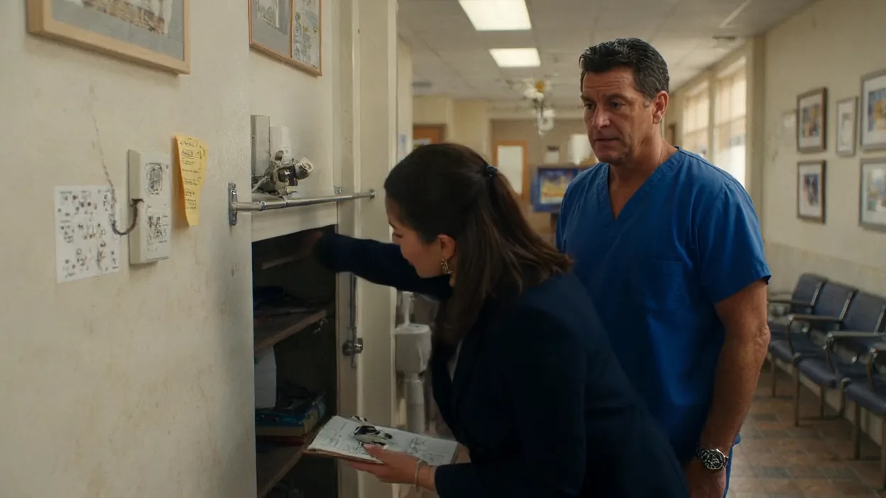 An assessor with a clipboard looks into an open wall cabinet of network equipment in a dental office hallway while a dentist watches.